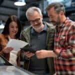 A group of three people—two men and one woman—are gathered around a car inside a well-lit auto dealership or repair shop. The woman, wearing a brown leather jacket, is holding a stack of papers, while the older man in a green jacket and glasses is smiling as he looks at a tablet being held by the man in a red plaid shirt. They appear to be engaged in a discussion, possibly reviewing vehicle information or documentation, with several cars visible in the background.