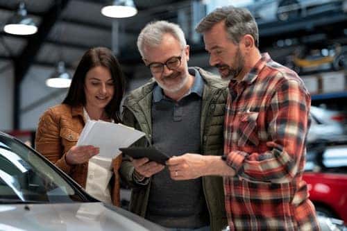 A group of three people—two men and one woman—are gathered around a car inside a well-lit auto dealership or repair shop. The woman, wearing a brown leather jacket, is holding a stack of papers, while the older man in a green jacket and glasses is smiling as he looks at a tablet being held by the man in a red plaid shirt. They appear to be engaged in a discussion, possibly reviewing vehicle information or documentation, with several cars visible in the background.