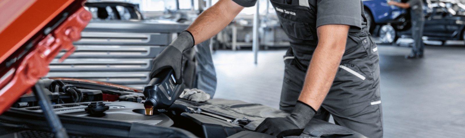 The image contains no captions. The image consists of a BMW Service Technician in gray coveralls leaning over the front of a Red BMW (model unknown) with the hood up looking at the engine compartment. The image shows the BMW Technician from the shoulders to the waist with one hand braced on the front of the car and the other hand is pouring in a quart of oil into the BMW. 