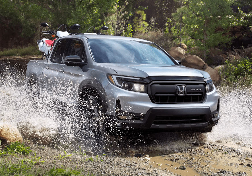 A silver 2024 Honda Ridgeline with two ATVs in the bed fords a large puddle on a dirt road