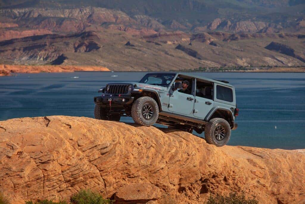 The 2025 Jeep Wrangler traversing red rocks in the desert with a body of water in the distance.