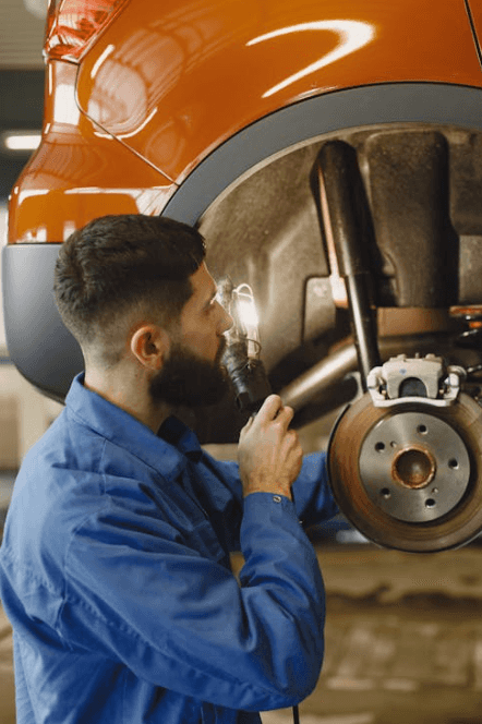 A bearded man in blue coveralls examines the front lefthand brake on a vehicle