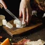 Woman hand cutting camembert on wooden board with other ingredients on table.