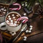 High angle view of two homemade hot chocolate mugs with marshmallows shot on rustic wooden Christmas table. A candy cane is inside one mug and another is placed directly on the table..