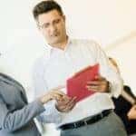 Man with red clipboard shopping for a pre-owned vehicle and speaking to a car saleswoman