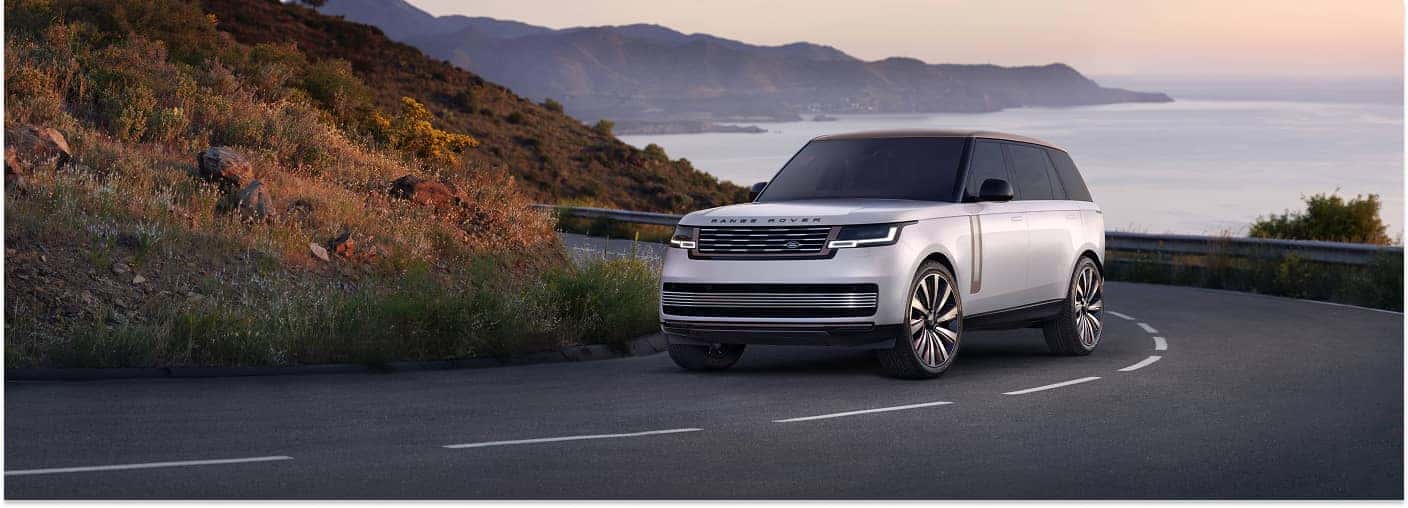 Front three-quarter view of a silver 2025 Range Rover SUV on a winding coastal road.