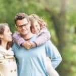 Young happy family of three having fun together outdoor. Pretty little daughter on her father back. Parents and girl look happy and smile. Happiness and harmony in family life. Family fun outside.