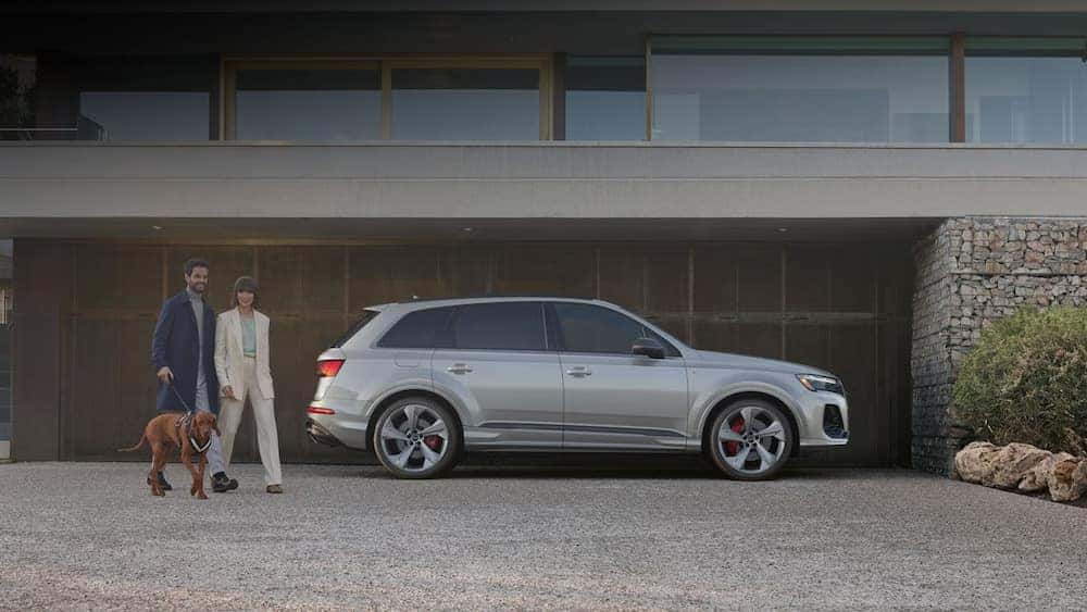 Two people walking a dog next to a gray 2025 Audi Q7 SUV parked in front of a modernist home