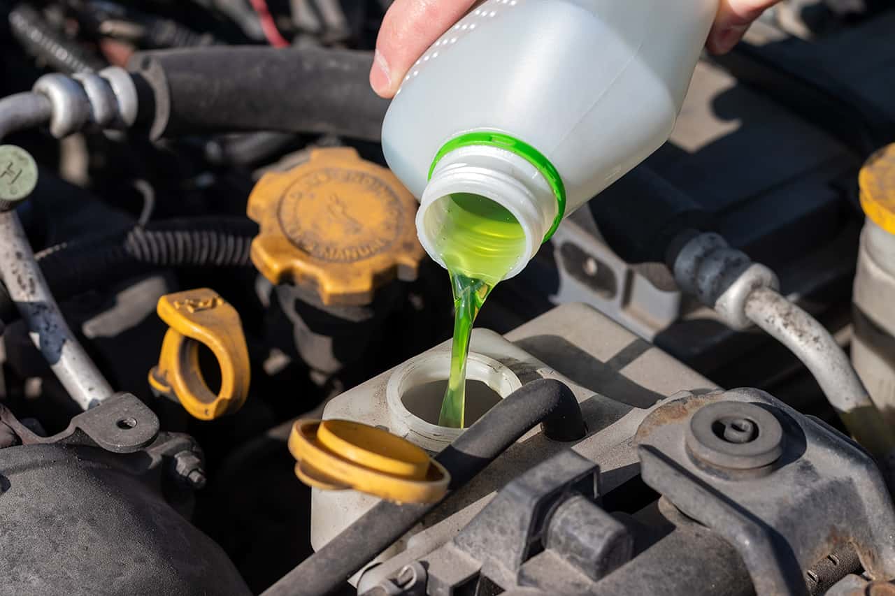Hand with bottle pouring antifreeze coolant into the expansion tank. Dusty details of a flat-four (boxer) car engine compartment under the open hood. Closeup view on a sunny day