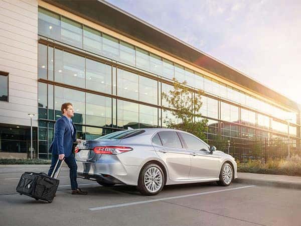 Person with a wheeled bacg walking up to a Toyota vehicle with modern building in the background